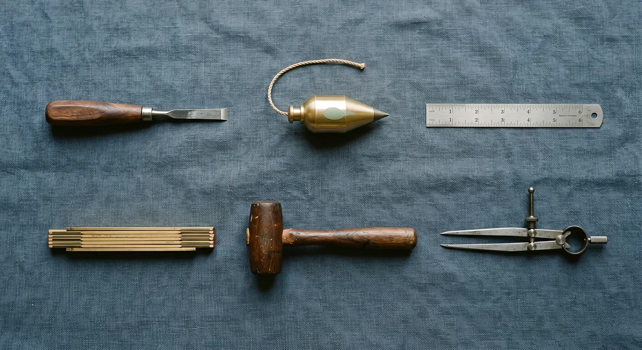 Six Tools — Craftsman's Bench Flat-Lay Flat-lay of six small handcraft tools — chisels, brass plumb bob, dividers, folding rule, mallet — on slate-blue linen, one with a sage-green grip